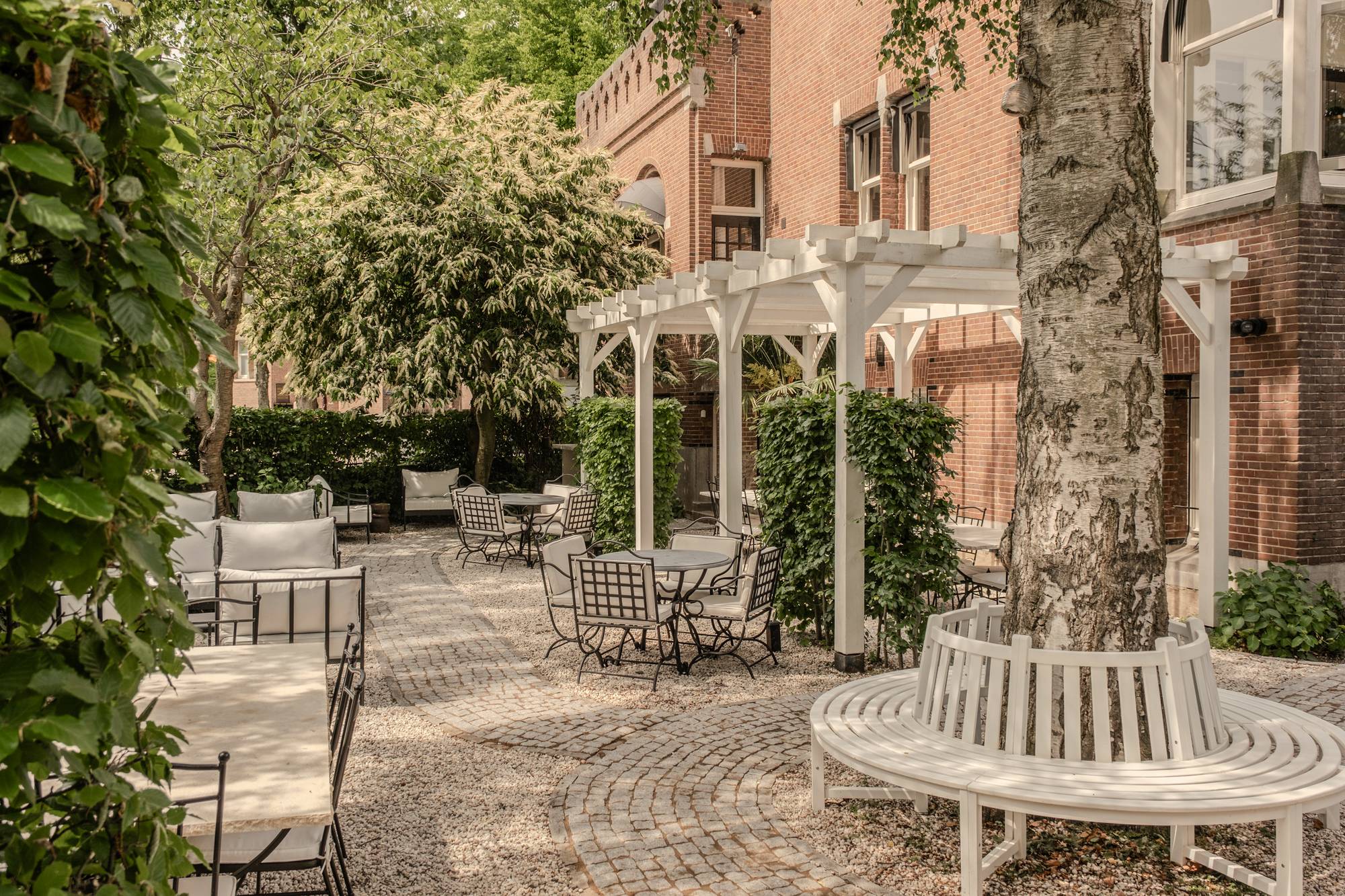 Garden at CreativeGrounds Emmalaan, Amsterdam Oud-Zuid, with a white pergola, a round tree bench, bistro tables and lounge sofas