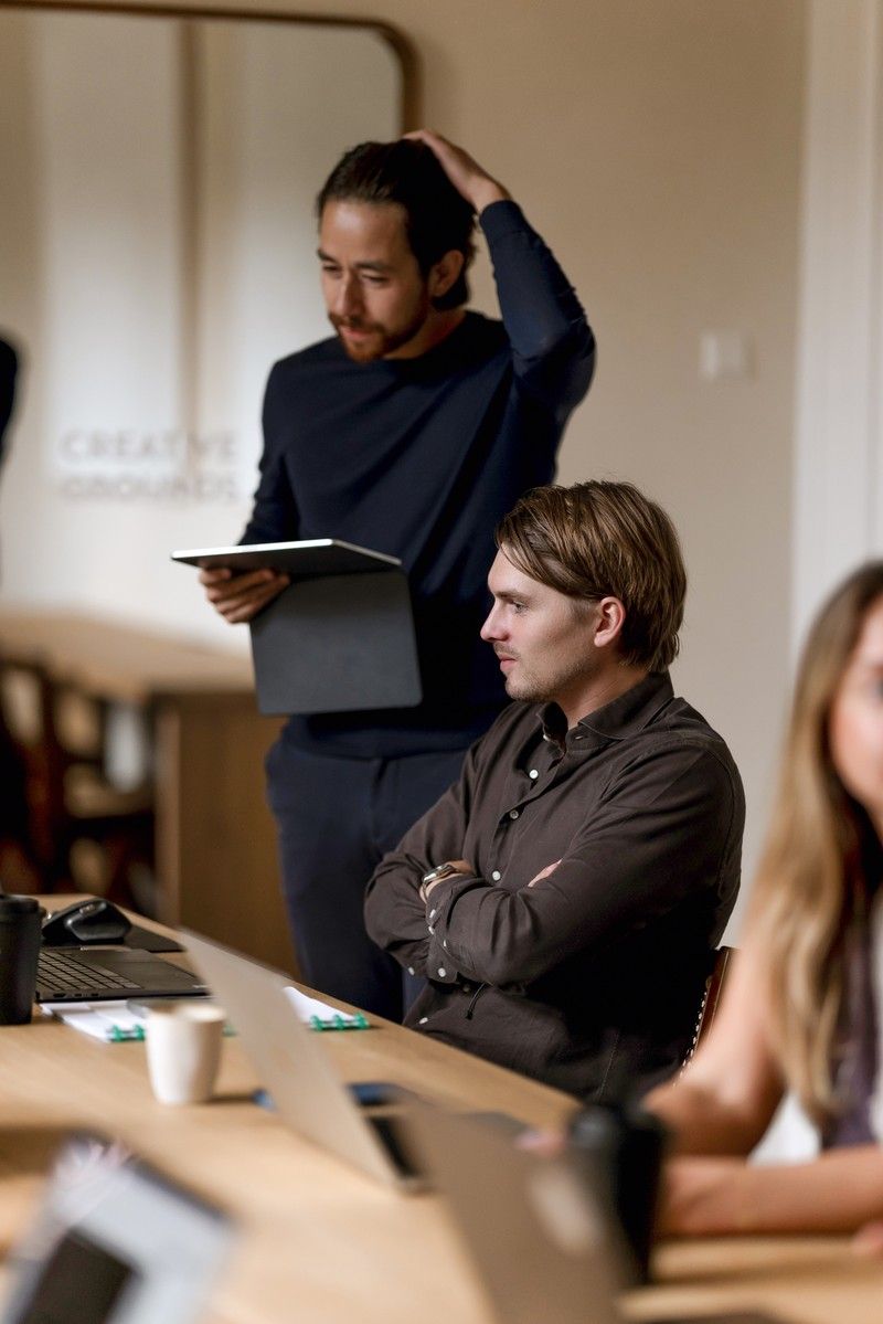 Two members at the coworking table at CreativeGrounds, one standing with a tablet, one seated with arms crossed