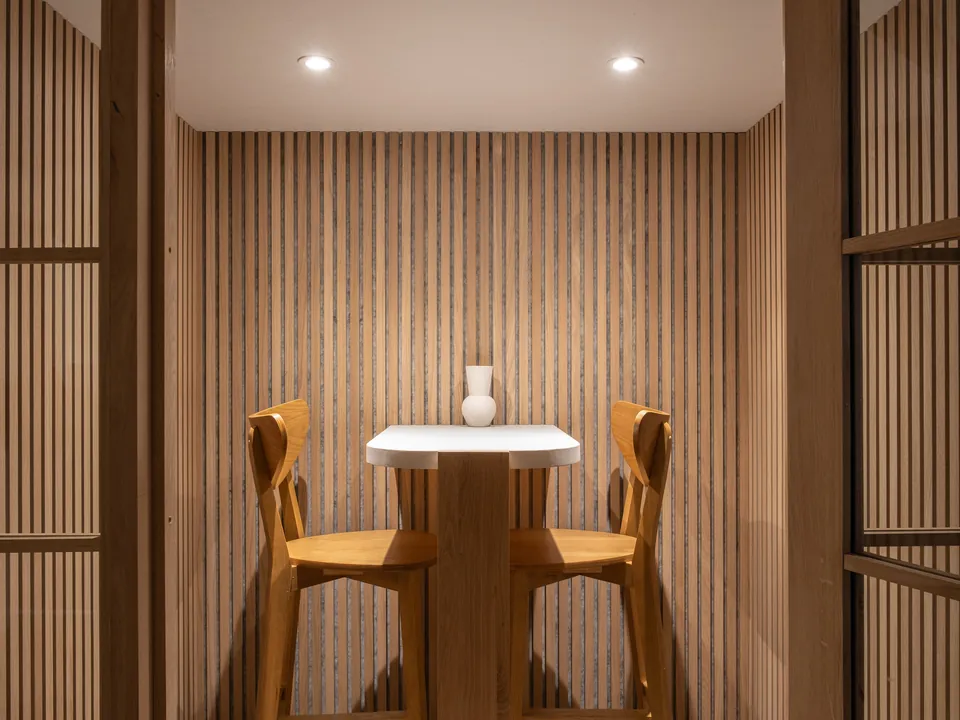 Wood-slatted phone booth with a small white table, two oak bar chairs and a white vase