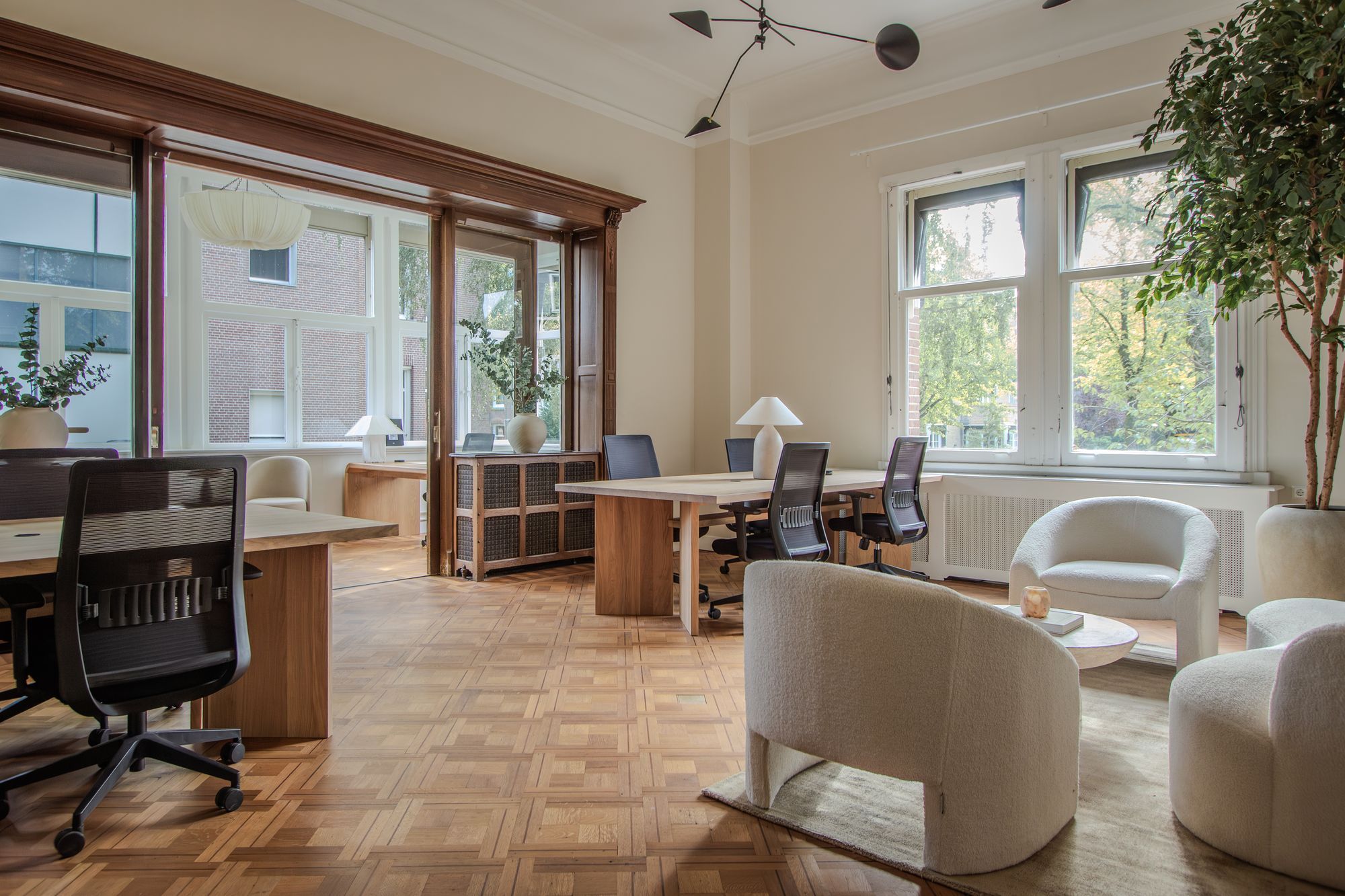 Bright private office with a wooden bay window, oak desks with black office chairs and cream armchairs around a low table in a CreativeGrounds villa in Amsterdam Oud-Zuid
